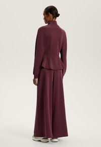 Burgundský blazer s peplum detailem, kombinovaný s širokými kalhotami. Hladký materiál, vysoký límec a volný střih, oblečený s robustními teniskami.