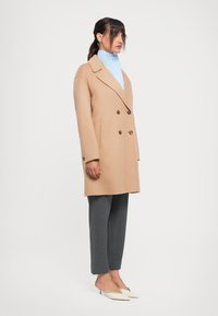 Donna in piedi con un cappotto doppiopetto beige, camicia azzurro chiaro con colletto alto, pantaloni grigi su misura e mules bianchi a punta con tacco basso, su sfondo bianco.