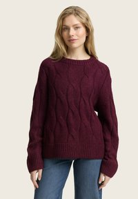 Bordeaux kabelgebreide trui met een oversized silhouet, geribbelde halslijn en manchetten, met een textuur in golfpatroon. Gemaakt in combinatie met blauwe jeans.