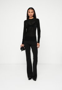 Črna dolga prosojna majica z teksturirano tkanino, v paru z črnimi oprijetimi hlačami in stiletto petami. Model drži majhno črno torbico.