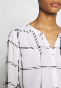 Vit blus med svart rutmönster, knappstängning och V-ringning. Materialet verkar vara lätt och har en slät textur.