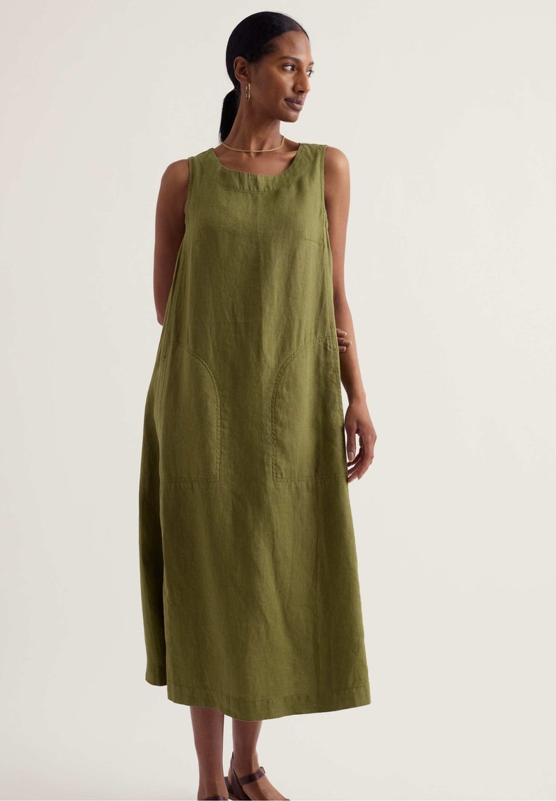 Femme aux cheveux foncés attachés en queue de cheval, portant une robe midi sans manches vert olive et des sandales marron, debout devant un fond clair uni.