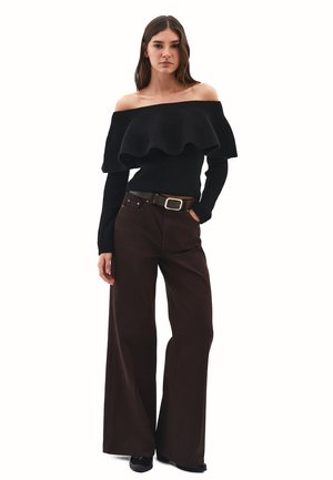 Schwarzer Schulterfreier Pullover mit gerafftem Ausschnitt, kombiniert mit weiten braunen Hosen und einem braunen Gürtel mit silberner Schnalle.