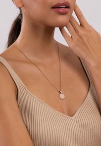 Goldene Kettenhalskette mit einem runden, blassen Anhänger und einem kleinen Akzentstein, getragen auf einem gerippten, neutralen Oberteil.