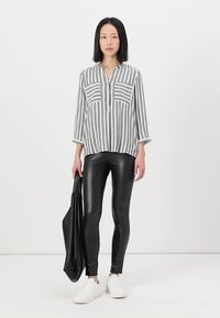 Gestreepte grijs-witte overhemden met lange mouwen, gecombineerd met strakke zwarte leggings en witte sneakers, terwijl ze een zwarte totebag vasthoudt.