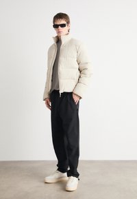 Jachetă puffer de culoare bej deschis, cu textură ribbed, guler înalt și închidere cu fermoar, asortată cu pantaloni negri lejeri și adidași bej.