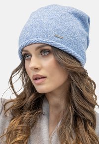 Bonnet en tricot bleu clair présentant un motif texturé, un rebord roulé et une étiquette en métal. Tissu doux, design décontracté et confortable.