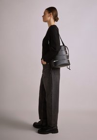 Sort læder bækken taske med sølv horisontale accenter, glat tekstur og justerbar rem, båret med en sort sweater og mørke jeans.