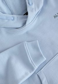 Sweat à capuche bleu clair avec poignets côtelés sur les manches, cordon visible et texte noir partiel sur la poitrine.