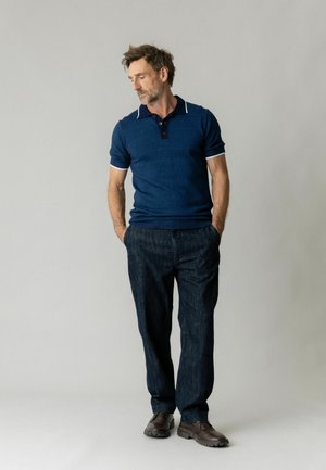 Uomo che indossa una polo blu a maniche corte e pantaloni scuri, in piedi con le mani nelle tasche, guarda verso il basso di lato.