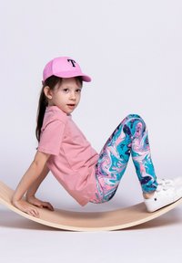 Różowa koszulka i różowa czapka z logo "T", w połączeniu z kolorowymi legginsami w wzór marmuru w odcieniach niebieskiego i różowego, białe sneakersy, na zakrzywionej drewnianej powierzchni.