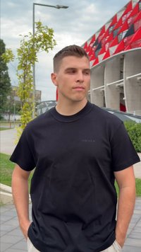 Jaunas vyras juodame SikSilk marškinėlyje stovi lauke šalia pastato su raudonais ir juodais geometriniais raštais, rankos kišenėse, žiūrėdamas į šoną.