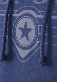 Sininen huppari, jossa on keskellä tähti graafinen kuvio, teksturoitu kangas ja kontrastivärinen nyöri. Graafisen suunnittelun takana on vaakasuuntaisia raitoja.