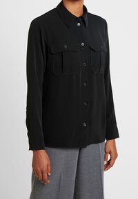 Camisa negra de manga larga con botones, cuello afilado y dos bolsillos delanteros con solapa, combinada con pantalones grises.