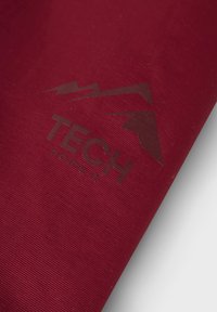 Detailný záber na tmavočervenú látku s logom hory a textom "TECH denim" vytlačeným na jej povrchu.