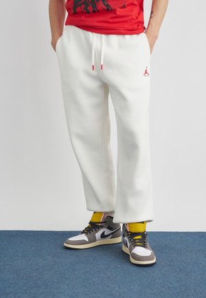 Persoană care poartă pantaloni de trening albi cu logo roșu, tricou roșu, șosete galbene și adidași negri, gri și albi, stând pe un covor albastru.