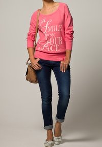 Roze sweatshirt met witte tekst, blauwe spijkerbroek en lichte schoenen. Vergezeld van een kleine bruin schoudertas. Casual outfit met opgerolde mouwen.