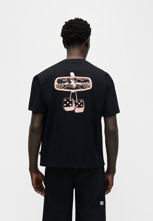 Fekete pólót viselő férfi, aki mögül látható a rózsaszín grafikus autótükör és lógó, bolyhos kockák.