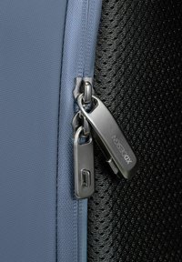 Sezione in tessuto azzurro chiaro con zip in metallo argentato, adiacente a un'area in rete nera testurizzata; presenta un piccolo logo sul tirante della zip.