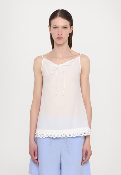 Camisole en lin blanc avec un décolleté en V, des bretelles ajustables et un détail de dentelle festonnée en bas. Associée à un pantalon bleu clair.
