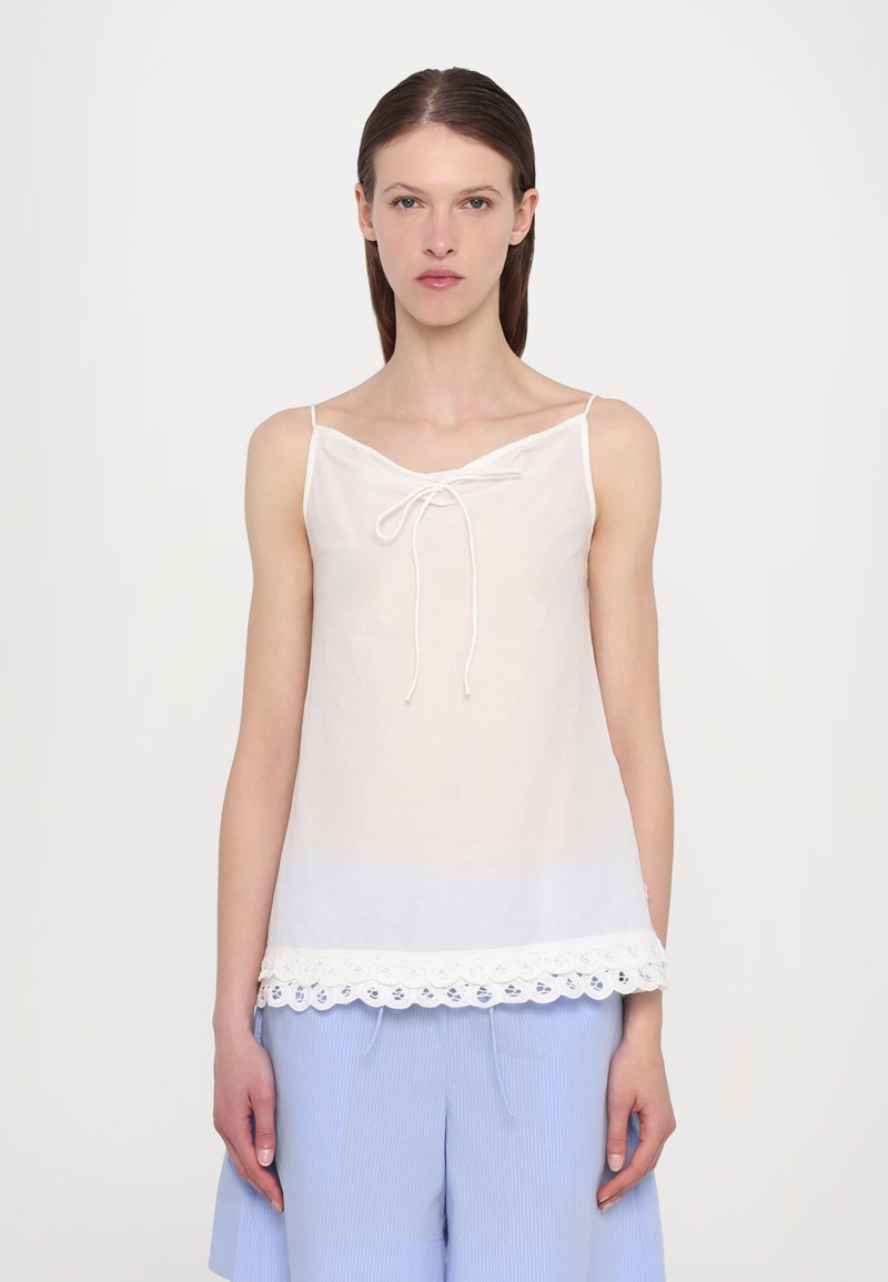 White linen camisole with a v-neckline, adjustable shoulder straps, and a scalloped lace hem detail. Paired with light blue pants.