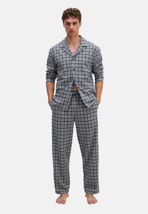 Homme debout pieds nus portant un ensemble de pyjama à carreaux noir et blanc assorti avec manches longues et pantalon à cordon, mains dans les poches, expression neutre.