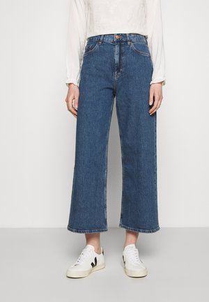 Person trägt hoch taillierte, weit geschnittene blaue Jeans, weiße langärmelige Bluse und weiße Sneakers und steht vor einem schlichten Hintergrund.