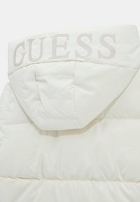 Valkoinen puffatakki, jossa on teksturoitu pinta, huppu, johon on kohokuvioitu sana "GUESS" vaalealla värillä, ja tikkipoliisi eristykseen.
