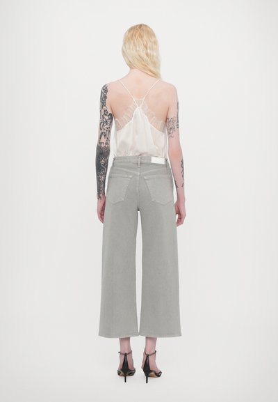 Jean large gris clair à taille haute, associé à un caraco crème translucide avec des bordures en dentelle et des fines bretelles. Talons noirs.