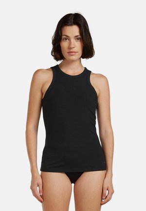 Mujer con cabello oscuro y corto que lleva una camiseta de tirantes negra acanalada y pantalones negros, de pie frente a un fondo blanco liso.