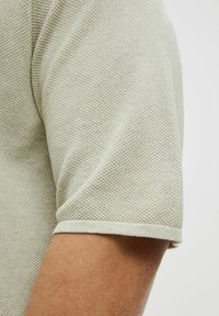 Chemise en tricot texturé beige clair à manches courtes, avec un ourlet arrondi et un poignet côtelé, présentant un motif tissé subtil.