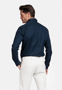 Chemise bleu marine à manches longues, avec des poignets boutonnés et un col pointu, portée avec un pantalon blanc, montrant une texture de tissu lisse.