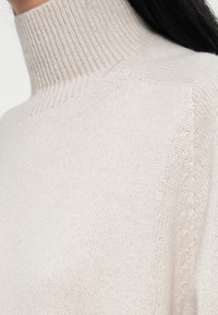 Close-up van een lichtbeige geribde coltrui die gedragen wordt, met zichtbare textuur en brei-details op de halslijn en schouder.