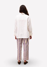 Eine Frau mit langen braunen Haaren steht mit dem Rücken zu uns, trägt ein lockeres weißes Hemd, vertikal gestreifte beige und lila Hosen sowie dunkelbraune Schuhe.