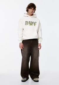 Witte fleece hoodie met "DWF"-grafiek in groen, gecombineerd met oversized bruine corduroy broek. Model draagt grijze schoenen met blauwe accenten.