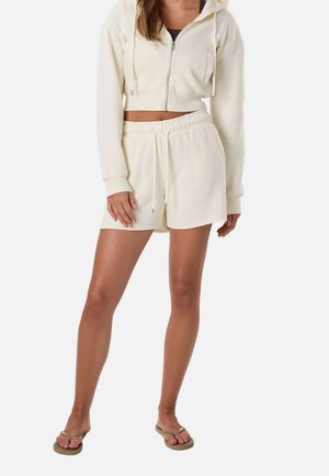 Femme portant un sweat à capuche court zippé de couleur crème, un short à cordon assorti et des tongs beiges, debout devant un fond blanc.