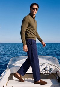 Homme portant des lunettes de soleil, un pull vert olive, un pantalon bleu marine et des mocassins marron, debout sur un bateau avec l'océan et un ciel dégagé en arrière-plan.