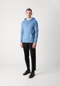 Sweatshirt azul claro com um bolso frontal, capuz com cordão e logo bordado. Combinado com calças pretas e sapatos pretos. Textura de tecido suave.