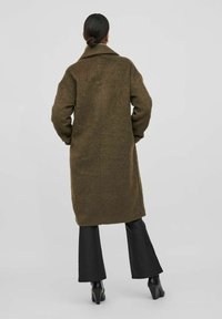 Vlněný kabát olivově zelené barvy s límcem, volným střihem, dlouhými rukávy a délkou do poloviny stehen, doplněný černými rozšířenými kalhotami.