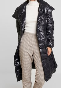 Abrigo de plumas negro brillante con un gran cuello, textura acolchada, cremallera frontal y cierres de botón a presión, combinado con pantalones de cuadros.