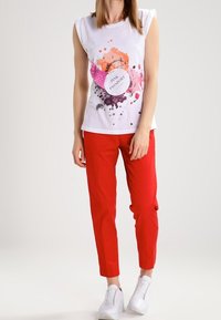 Biała, bezrękawnikowa koszulka z kolorowym nadrukiem w kropki i napisem "PINK PASSPORT", w połączeniu z czerwonymi spodniami slim-fit i białymi trampkami.