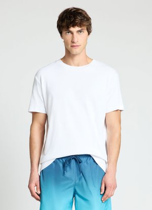 Junger Mann mit heller Haut und kurzen braunen Haaren, der ein schlichtes weißes T-Shirt und blaue Shorts mit Farbverlauf und Kordelzug trägt, steht vor einem weißen Hintergrund.