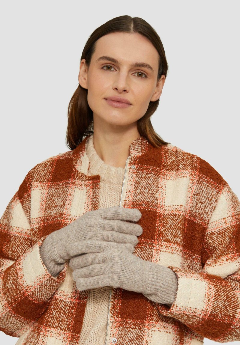 Grijze wollen handschoenen met geribde manchet. Model draagt een geblokte oranje en crèmekleurige jas over een beige trui. Eenvoudig en functioneel ontwerp.