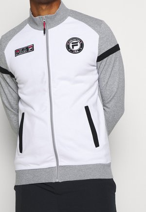 Mann trägt eine weiße und graue Jacke mit Reißverschluss, schwarzen Streifen an den Ärmeln und FILA-Logos auf der Brust.