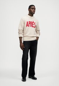 Béžový pletený svetr s červeným nápisem "APRÈS", kulatý výstřih, žebrované manžety, doplněný černými džínami a černými botami. Jednoduchý, strukturovaný design.