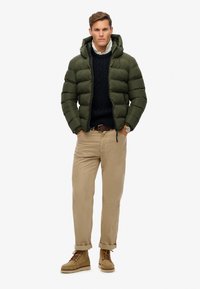 Giacca a vento verde oliva, con cappuccio e design trapuntato. Indossata sopra un maglione blu navy a trecce, abbinata a pantaloni chinos beige e stivali marroni.