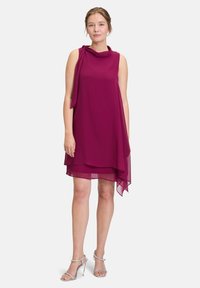 Robe sans manches magenta avec un décolleté drapé, un ourlet asymétrique et un superposé transparent. Portée avec des sandales à talons hauts argentées.