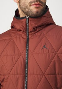 Red quilted jacket featuring a black zipper and a small black logo on the chest. The material has a smooth texture with a hooded design.