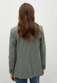 Veste à carreaux verts et blancs avec une coupe ajustée, une fente simple à l'arrière et des manches à boutons. Portée sur un jean en denim.
