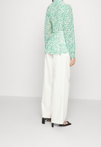 Grün-weiße gemusterte Bluse mit langen Ärmeln und Peplum-Design, kombiniert mit weißen weit geschnittenen Hosen und schwarzen Absatzsandalen.